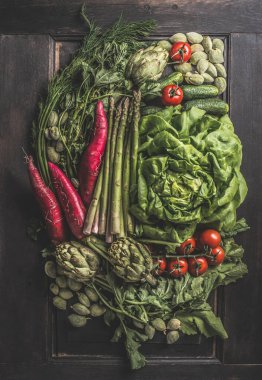 Bir sürü farklı sebze. Esmer arka planda taze ekilmiş enginar, salata, domates, kuşkonmaz, turp, yeşil mandalina, salatalık ve lezzetli yemekler için bitkilerle birlikte kompozisyon. Üst görünüm