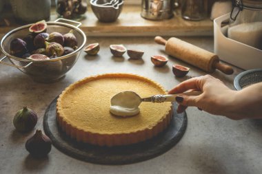 Kadın eli bisküvi kekine krem sürerken beton mutfak masasında dilimlenmiş incir ve incir, ahşap oklama iğnesi ve pişirme aletleriyle. Evde taze malzemelerle pişirmek. Ön görünüm.