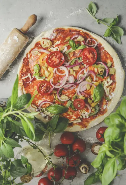 Pizza preparation with salami, tomatoes, mushrooms, onion, basil, cheese , fresh herbs and rolling pin on grey table. Cooking traditional Italian food at home with fresh ingredients. Top view