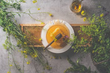 Balpeteğinin üzerine bal kaşığı konmuş taze bal. Etrafı yabani çiçekler ve mutfak gereçleriyle çevrili. Doğal, organik gıda konsepti. Koyu beton arka plan. Üst görünüm