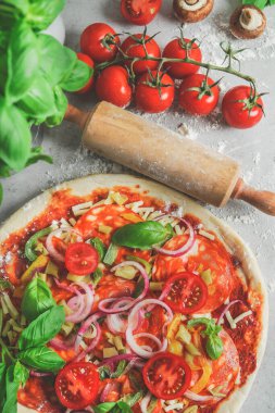 Domatesli, salamlı, soğanlı ve fesleğenli ev yapımı pizza. Mutfak masasında un, tahta raptiye, fesleğen ve malzemelerle. Geleneksel İtalyan yemekleri pişirmek. Üst görünüm.