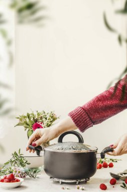 Kadın elleri mutfak masasında siyah tencereyi tutarken, arka planda çiçekler ve ahududular vardı. Evde yemek pişirmek. Ön görünüm.