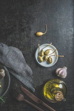 Koyu beton mutfak masasında kapari, zeytin yağı, sarımsak, çatal bıçak ve masa örtüsü. Akdeniz yemeği aperatifi. İtalyan yemeği turşusu. Kopyalama alanı olan üst görünüm.