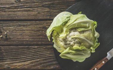 Kırsal ahşap mutfak masasında bıçakla siyah kesme tahtasındaki bütün ham karnabaharı kapat. Sağlıklı pişmemiş sebze. Yemek hazırlığı. Üst görünüm.