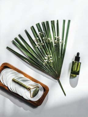 Masaj rulosuyla cilt bakımı, pipetli yeşil cam şişe serumu ve güneş ışığı ve gölgeli beyaz masada palmiye yaprağı. Sağlıklı güzellik ve besleyici ürünler. Üst görünüm.