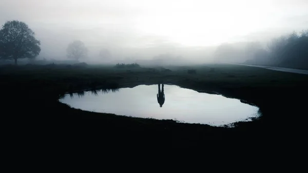 A haunted concept of the reflection of a man who isn\'t there. Standing by a pond. On a spooky mist evening in the countryside