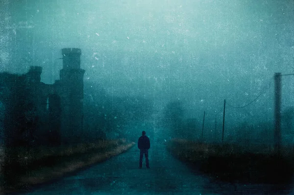 一个男人站在一个被废弃的废墟旁边的多雾的乡间道路上 用恐怖的咕噜声编辑 图库照片