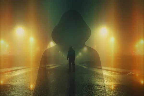 A double exposure of a mysterious hooded figure. Looking down an empty city street. On a spooky foggy winters night. With blurred abstract glowing lights.
