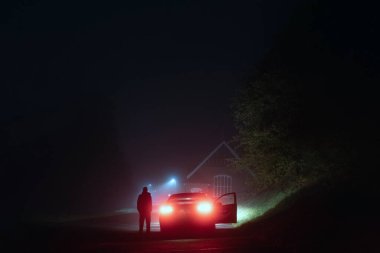 Sisli bir gecede ürkütücü, boş bir yolda bir arabanın yanında duran gizemli bir adam. Sokak lambalarının gölgesinde.
