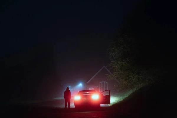 在一个雾蒙蒙的夜晚 一个神秘的男人站在一条令人毛骨悚然的空旷道路上的一辆汽车旁边 被街灯勾勒的轮廓 图库图片