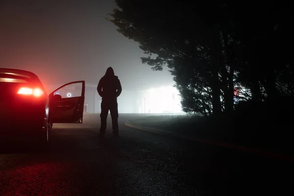 一个神秘可怕的戴着头巾的人影在一个阴云密布的令人毛骨悚然的夜晚 站在一辆停放着的汽车旁边 车后的灯光闪烁着建筑物的光芒 图库图片