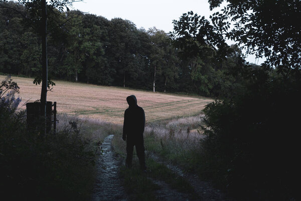 Странная, загадочная, страшная фигура в капюшоне. Стоя на фермерской дорожке в сельской местности. Окруженный лесом в мрачный вечер.
