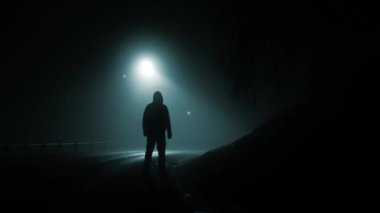 Ürkütücü bir kır yolunda gökyüzünde hareket eden gizemli doğaüstü UFO ışıklarını izleyen kapüşonlu bir figür.. 