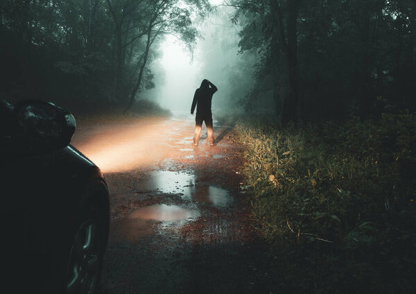 Ужасная концепция силуэта жуткой фигуры в капюшоне, стоящей в жутком лесу в туманный вечер. Выделенные фары автомобиля на грязной дорожке.