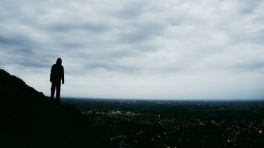 Kapüşonlu gizemli bir figür, tepede duruyor ve akşamları parlayan sokak ışıklarıyla bir kasabaya bakıyor..