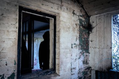 Ürkütücü bir korku konsepti. Harabe bir evde. Camdan süzülen ürkütücü, koyu renkli kapüşonlu bir siluetle.