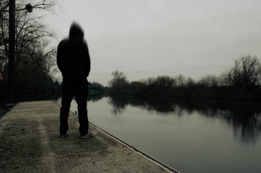 Akıl sağlığı konsepti. Kapüşonlu gizemli bir figür, kasvetli bir kış gününde, bulanık bir kurguyla nehri seyrediyor..