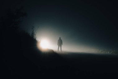 Ürkütücü kapüşonlu bir figür. Bir köy yolunda siluetlendi. Gizemli bir sisli kış yolunda geceleyin