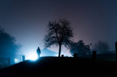 Parkta dikilen tüyler ürpertici kukuletalı bir figür. Sokak lambaları tarafından siluetlendi. Ürkütücü bir kış gecesinde