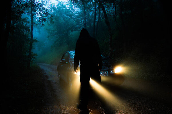 Ужасная концепция фигуры в капюшоне. Стоя перед машиной с включёнными фарами. В туманном туманном лесу в сумерках.