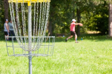 Disk golf oyuncusu parkta uçan bir disk atıyor, odak noktasında zincir sepet.