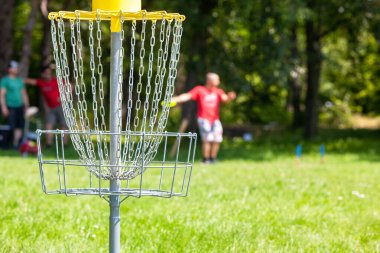 Disk golf oyuncusu parkta uçan bir disk atıyor, odak noktasında zincir sepet.