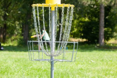 Disk golf oyuncusu parkta uçan bir disk atıyor, odak noktasında zincir sepet.