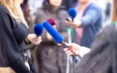 Arkaplanda haber, basın toplantısı veya medya karmaşası, mikrofon tutan kadın muhabir, diğer gazeteciler ve kamera operatörü