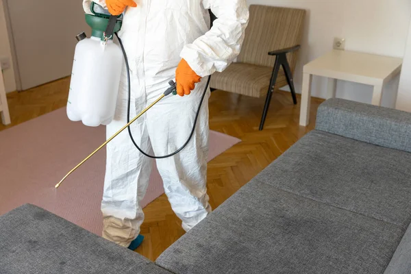 Person in protective suit with decontamination sprayer bottle disinfecting household and furniture. Pest control concept.