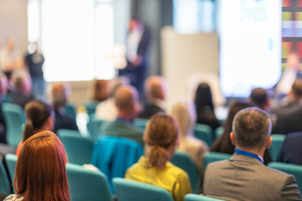 Профессиональная конференция или семинар, фокусируясь на аудитории, сидящей в ряд, слушая спикера, который размыт в фоновом режиме