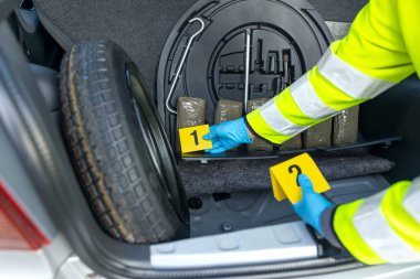 Polis memuru, bir arabanın bagajında bulunan uyuşturucu paketlerini kanıt işaretleriyle araştırıyor.