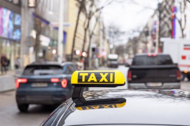 Şehir caddesinde odak noktasında bir taksi tabelası ve kalabalık bir şehir ortamında araçlar yer alıyor.
