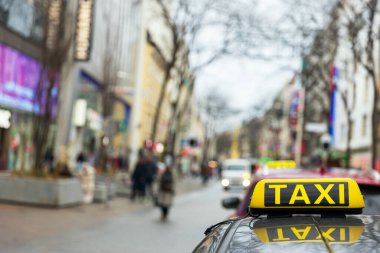 Şehir merkezinde yayalar ve araçlarla dolu bir şehir caddesine odaklanan taksi tabelası