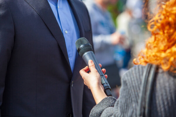 A journalist with curly red hair holding a microphone interviews a formally dressed individual outdoors during a public event