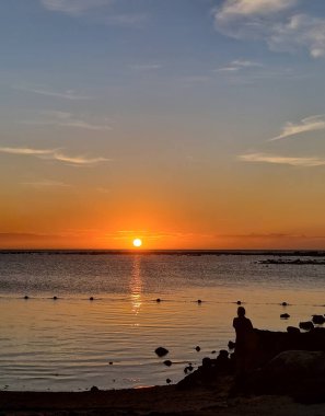 Huzurlu bir günbatımı, okyanus üzerinde sıcak bir parıltı yayar, ufka karşı siluet oluşturan bir insan, huzuru ve yansımayı çağrıştırır..