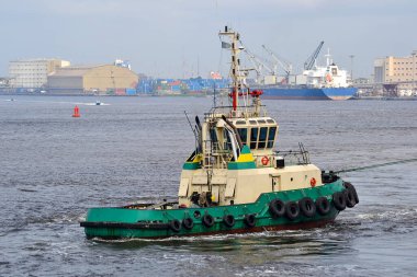 Römorkör limanda yelken açıyor. Büyük bir kargo gemisine yardım ediyor. Endüstriyel limanda denizcilik faaliyetleri var.