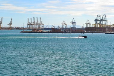 Valencia, İspanya 'da canlı bir limanda büyük vinçler ve sürat teknesi bulunur. Açık gökyüzü ve uzak şehir manzarası deniz aktivitesini arttırır..
