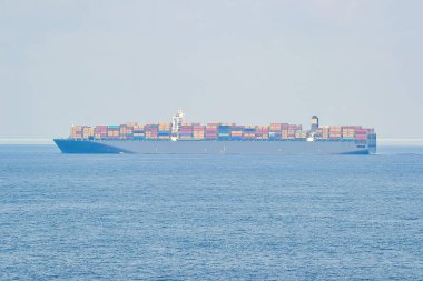 Renkli nakliye konteynırlarıyla dolu büyük bir konteyner gemisi sakin bir denizde yol alıyor. Gemi berrak, soluk mavi bir gökyüzüne karşı dizilmiştir..