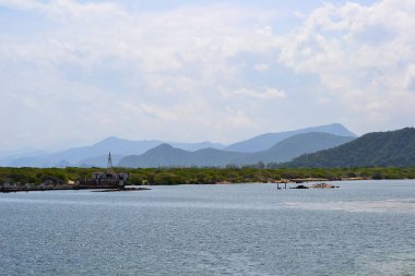 Arkasında yemyeşil tepeler ve dağlar olan sakin bir su kütlesi. Dar bir kara şeridinde kulesi olan küçük bir köy yapısı duruyor..