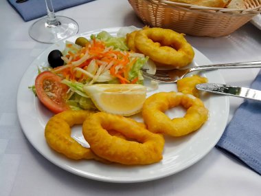 Bir tabak kızarmış kalamar, salata, limon ve çatal bıçak takımı. Arka planda beyaz bir masa örtüsünün üzerinde bir sepet ekmek ve bir bardak şarap var..