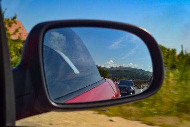 A car's side mirror reflects a scenic landscape with rolling hills and a clear blue sky. A line of cars is visible, suggesting a travel scene in a rural area. clipart