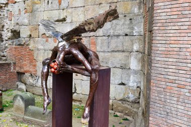 Kanatlı bir figürün modern metalik heykeli, antik taş ve tuğla duvarlarla çelişen bir heykel üzerinde duruyor..