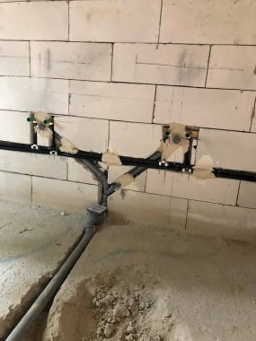 Borularla korunmasız tesisat sistemi ve hafif tuğladan bir duvara monte edilmiş. Toprak ve molozlarla dolu tamamlanmamış zemin, devam eden inşaat çalışmalarını gösteriyor..