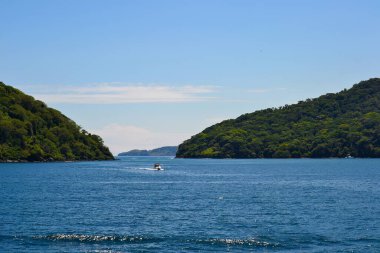 A small boat travels through a narrow channel flanked by green hills, with deep blue water reflecting the clear sky in a tropical or subtropical setting. clipart