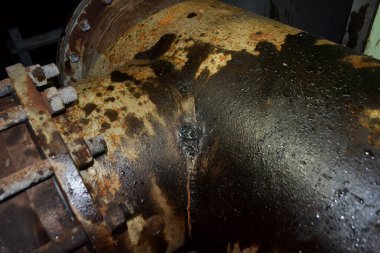 Büyük bir endüstriyel boru gözle görülür çatlaklar ve aşınmayla birlikte önemli bir pas ve yağ sızıntısı gösteriyor. Sürgüler ve kenarlar sanayi ortamını vurguluyor.