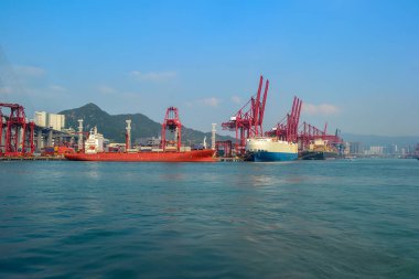 Büyük kargo gemileri Hong Kong limanında, yüksek kırmızı vinçlerle çevrili. Arka planda dağlık bir manzara ve kentsel yapılar görülebilir..