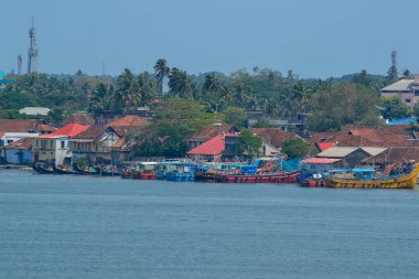 Hindistan 'daki bir sahil sahnesinde canlı balıkçı tekneleri, kırmızı çatılı binalar, ve gelişmiş bir bölgeyi gösteren iletişim kuleleri ile bereketli palmiye ağaçları yer almaktadır..