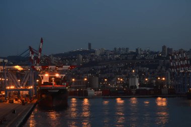 İsrail, Hayfa 'da, alacakaranlıkta canlı bir liman sahnesi. Aydınlatılmış turnalar ve bir kargo gemisi görülebiliyor. Arka planda da Baha 'i Bahçeleri var..