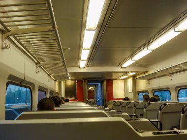 Görüntü banliyö treninin içini gösteriyor. Sıralı koltuklar, üstteki bagaj rafları ve floresan aydınlatma. Yolcular oturuyorlar, bazıları bulanık manzaraya bakarak trenin hareket ettiğini gösteriyor. Tasarımına göre...