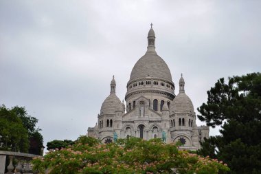 Fransa 'nın başkenti Paris' teki Sacre Coeur Bazilikası 'nda beyaz kubbeler, karmaşık detaylar ve bulutlu bir gökyüzü altında yeşillik ve çiçek açan ağaçlar yer almaktadır..